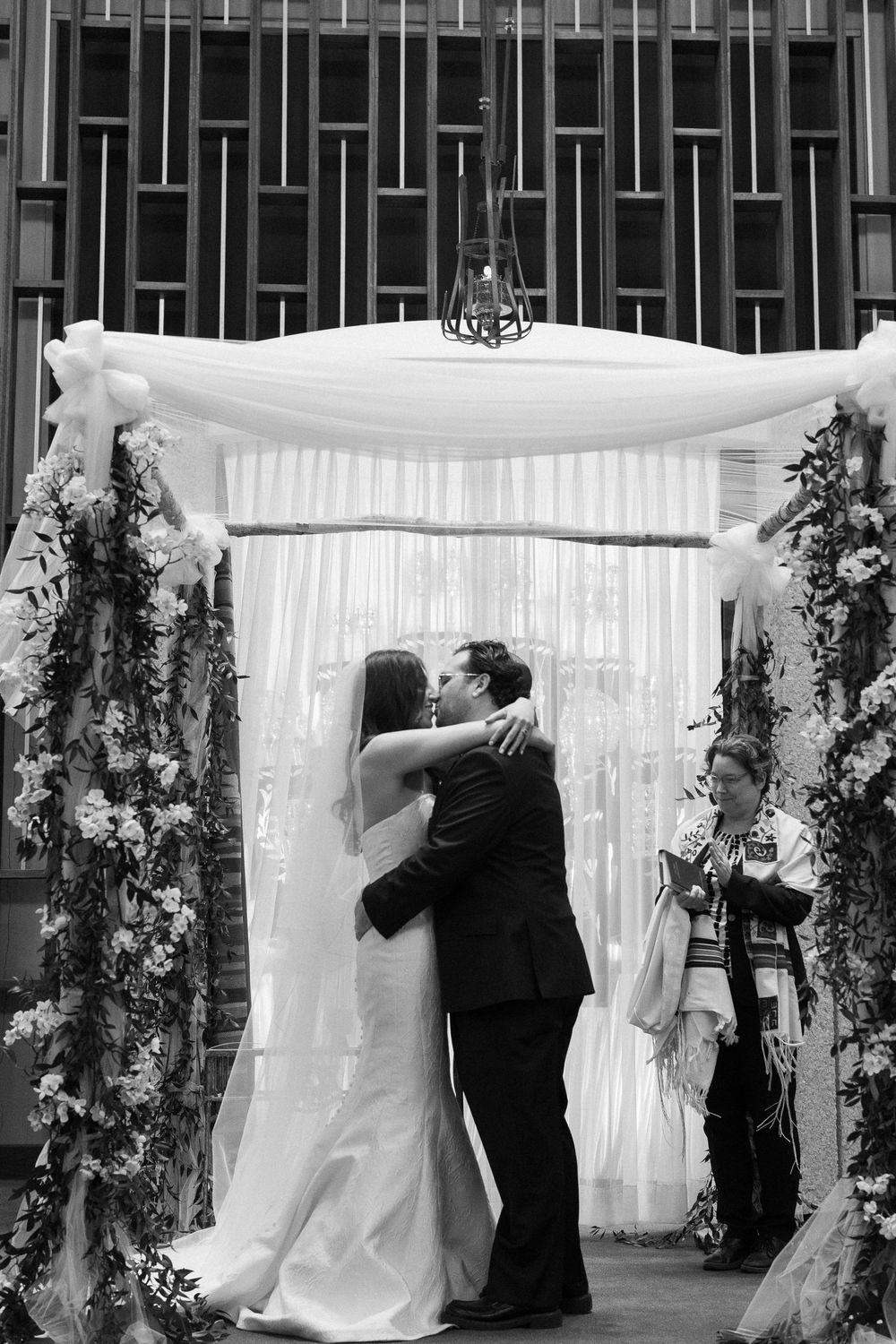 Black and white photo of bride and groom sharing first kiss under floral chuppah at Jewish wedding ceremony.