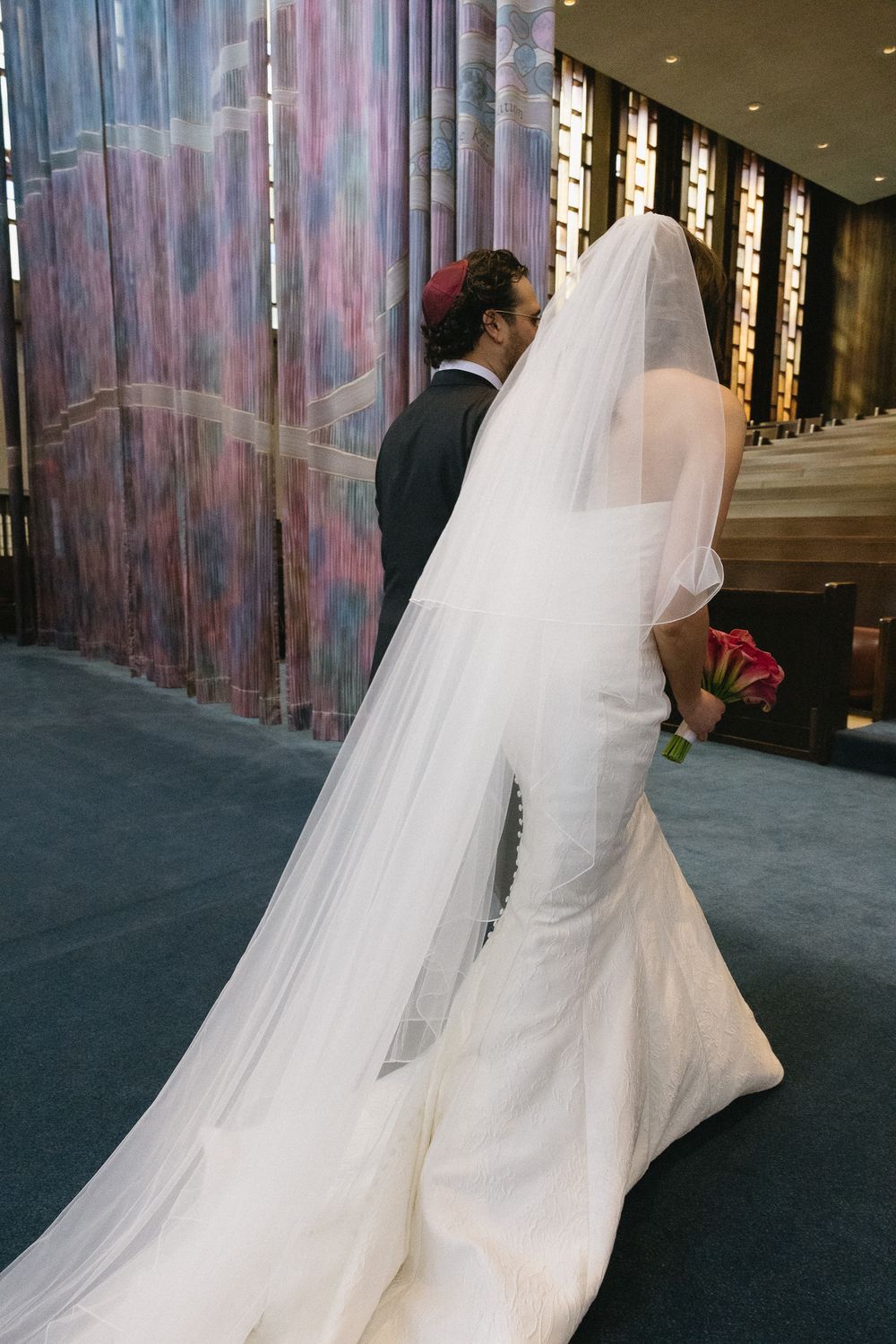 Bride in white gown with long veil walks with groom in black suit during wedding ceremony in elegant venue.
