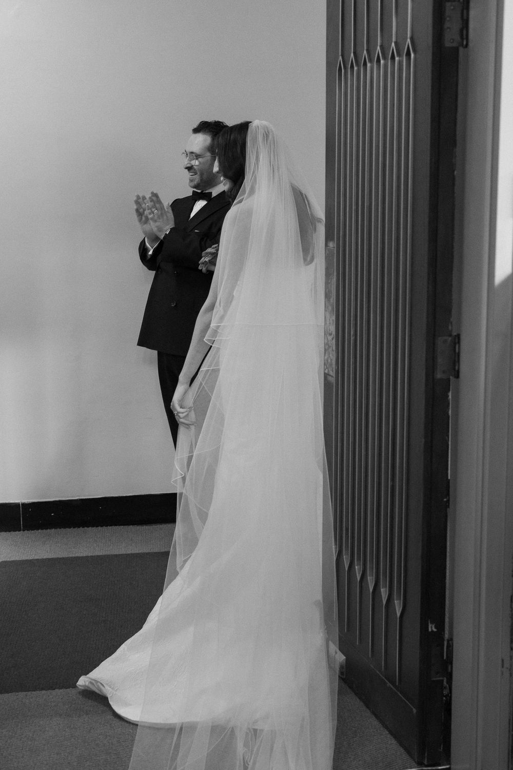 Black and white photo of a bride in a flowing veil and groom in a tuxedo standing by a door before their wedding ceremony.