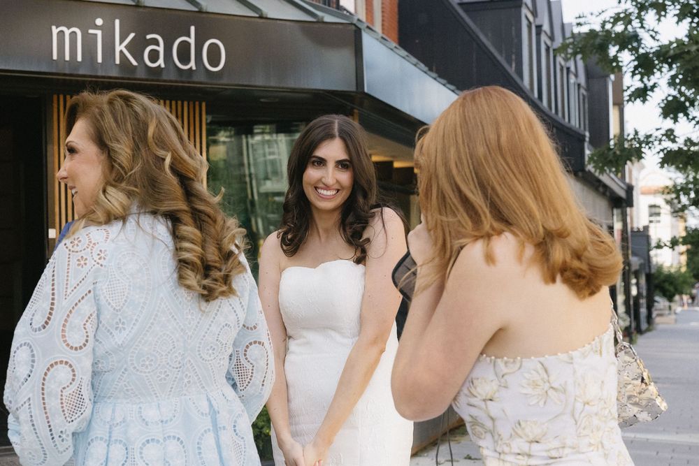 Bride in white strapless gown laughing with bridesmaids outside Mikado bridal boutique on a sunny day.