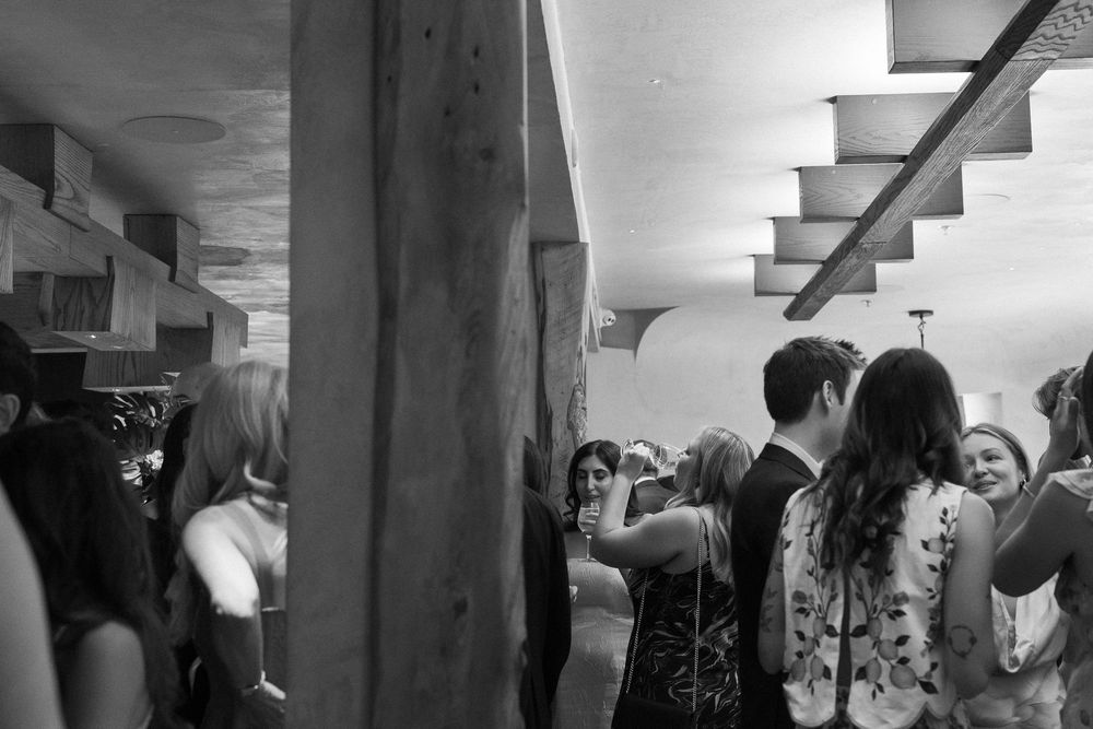 Black and white photo of a crowded indoor party with guests socializing near concrete pillars and a staircase.