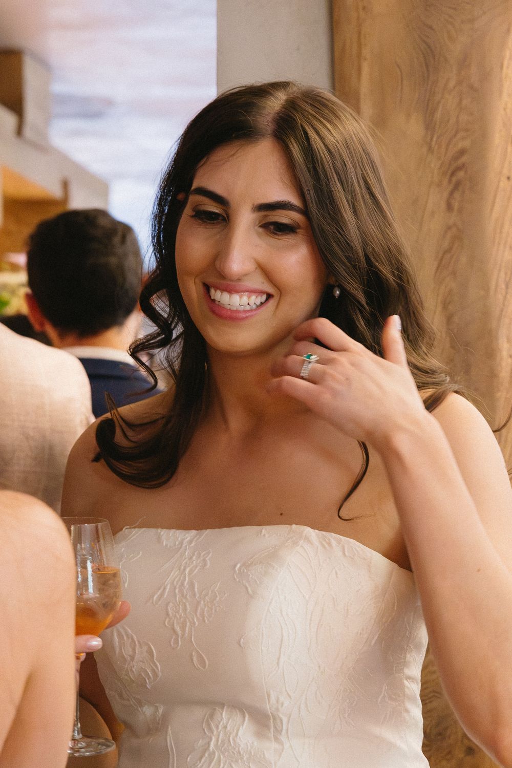 Smiling bride in strapless white gown holding champagne, showing off her engagement ring at a wedding reception.