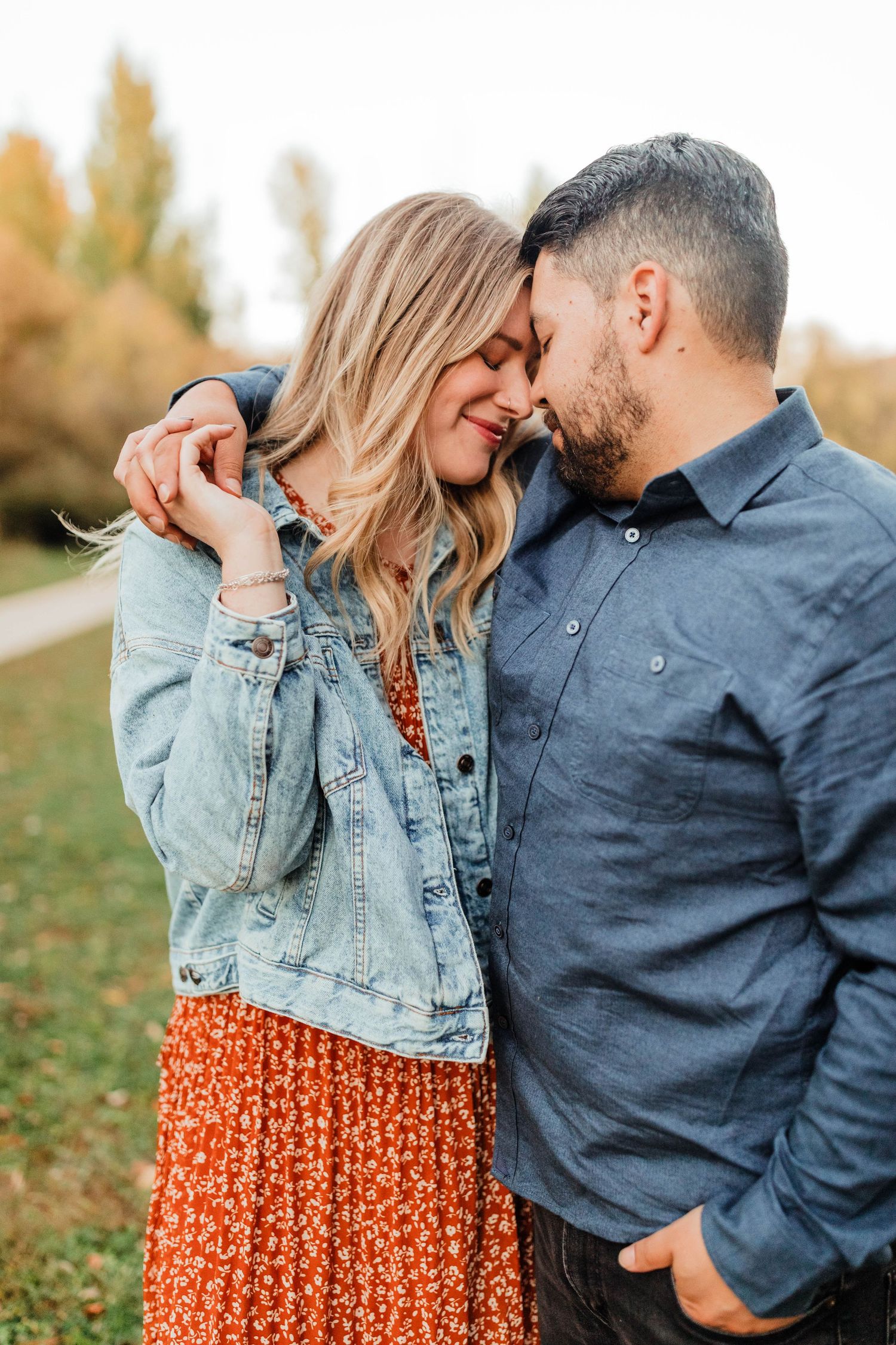 Couple embracing outdoors wearing denim jacket and navy shirt against autumn backdrop.