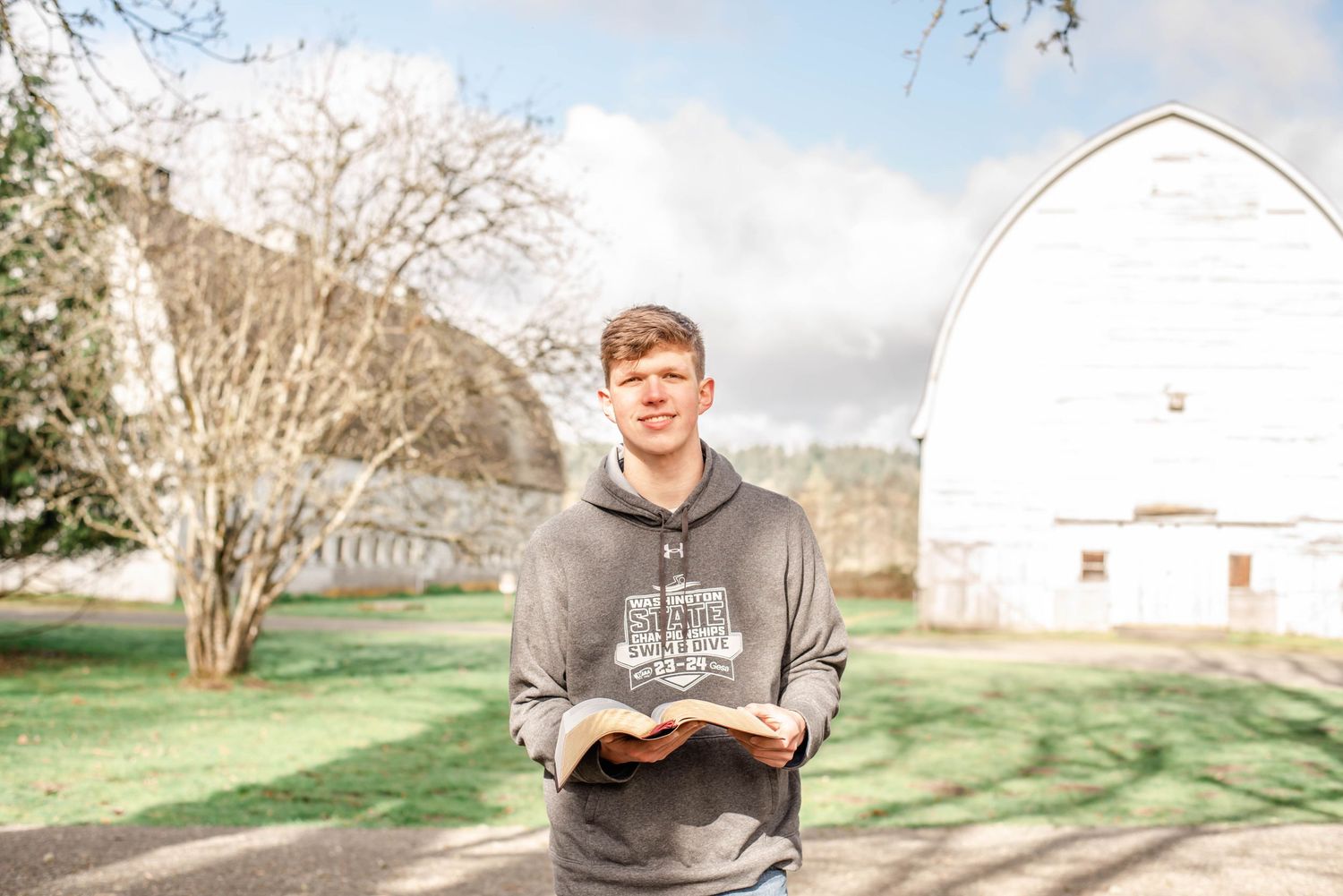 Person in grey hoodie reading book outdoors with white barn in background.
