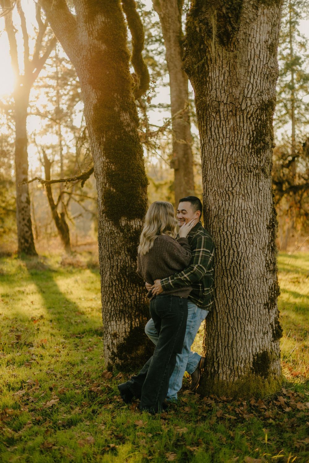A playful moment captured in a sunlit forest as a couple leans against trees during golden hour.