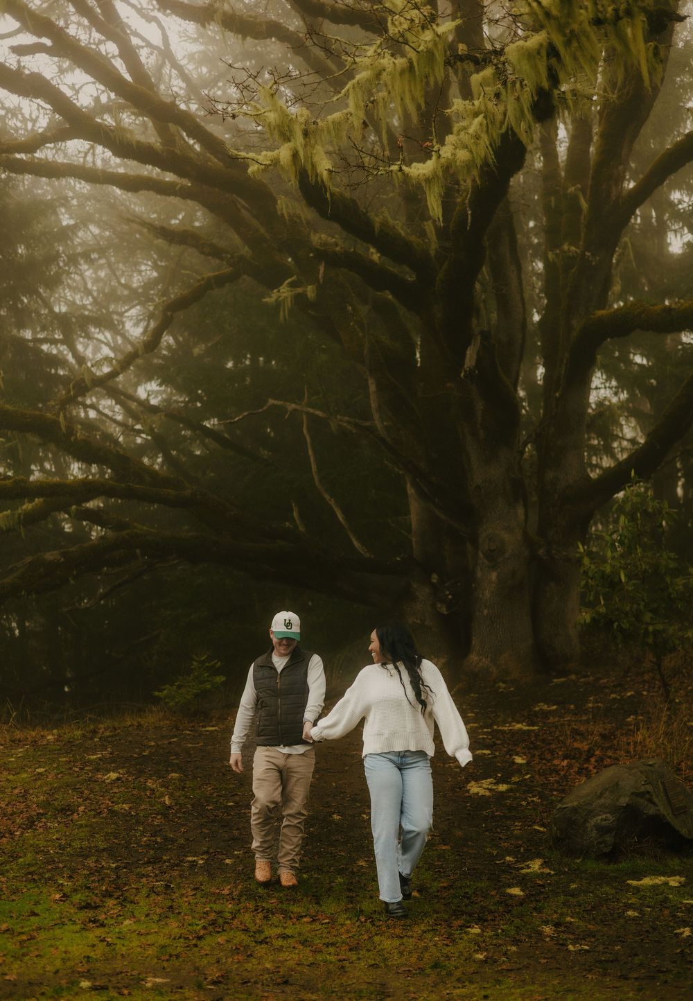 Two people walking hand in hand through a misty forest with moss-covered trees creating a moody atmosphere.