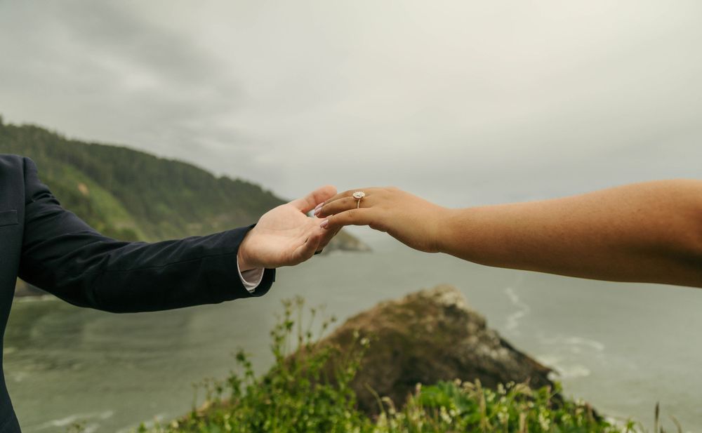 Couple holding hands at a scenic coastal overlook with misty ocean and green hills in the background.