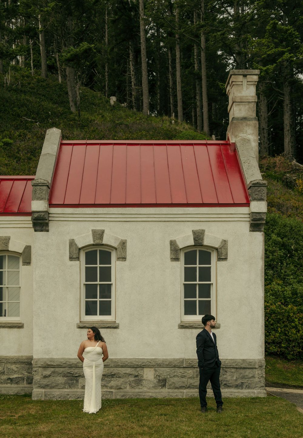 Bride in white gown and groom in navy suit stand apart before a stone building with red roof, surrounded by lush trees.