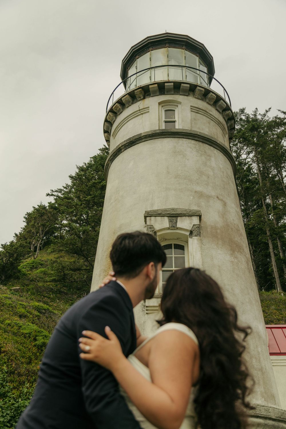 A couple shares a romantic kiss in front of a historic lighthouse surrounded by lush green trees on a cloudy day.