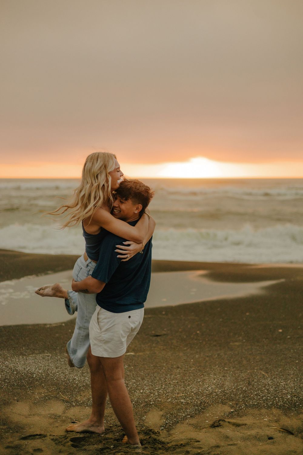Couple embraces romantically on a sandy beach at sunset with ocean waves in the background.