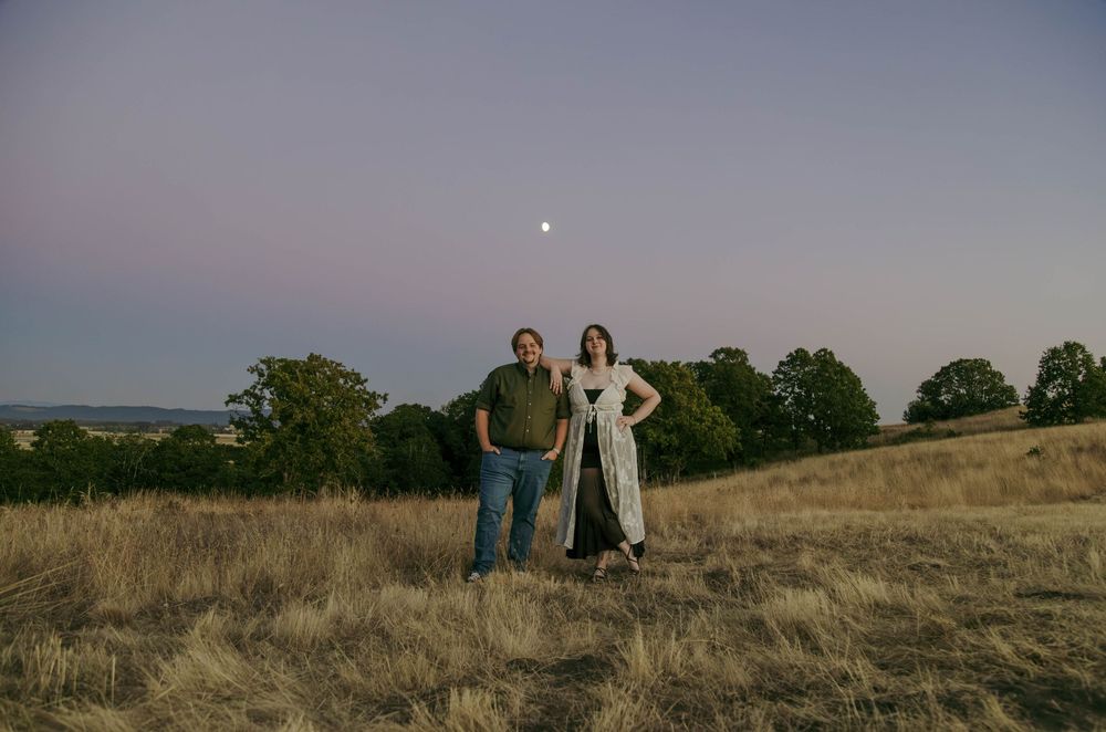 Couple standing in golden field at dusk with moon visible in purple sky and trees in background.