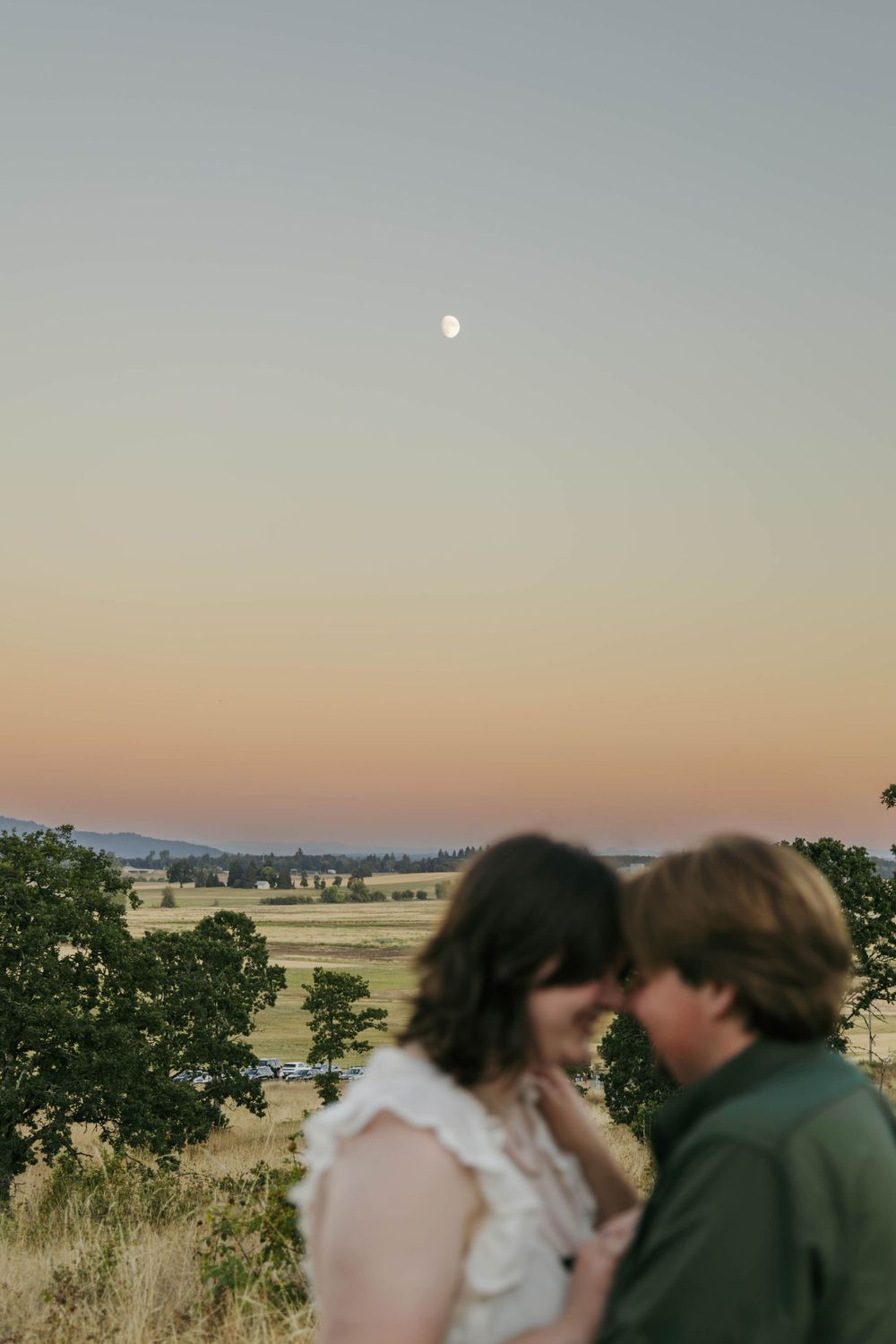 Couple shares romantic moment at sunset with full moon rising over golden fields and trees in background.