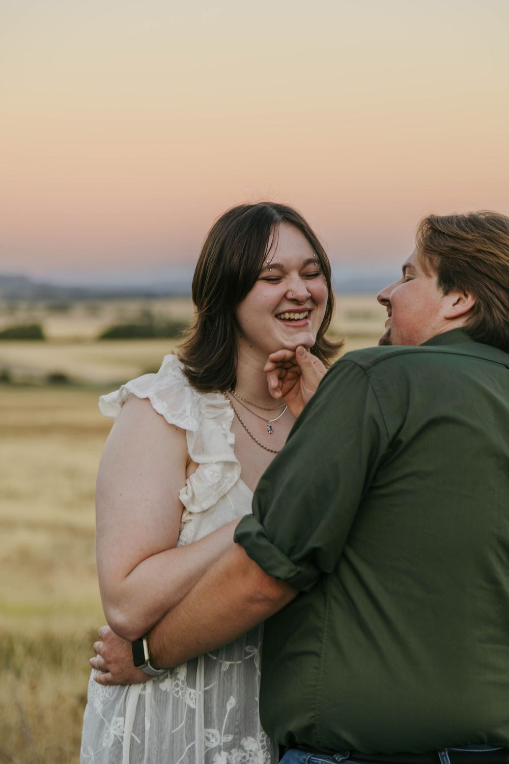Two women laughing and embracing at sunset in a golden field, one in white dress, other in green shirt.