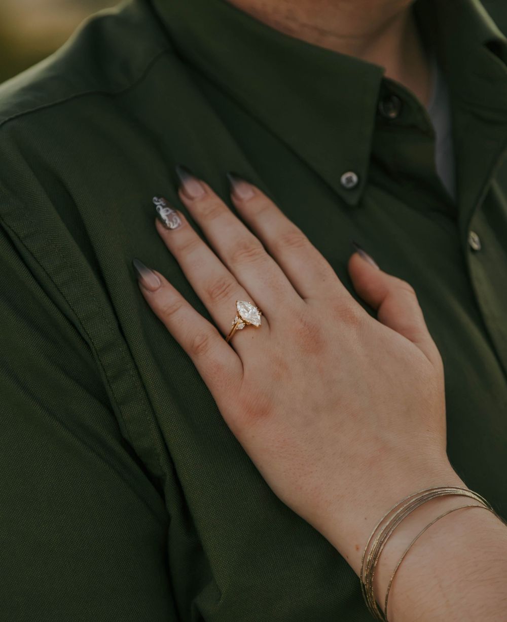 Close-up of a hand with dark nails wearing a gold engagement ring, resting on a green button-up shirt.
