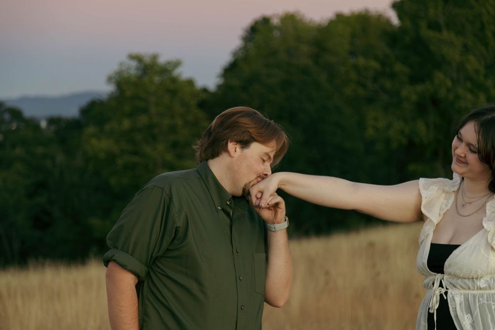 A man kisses a woman's hand outdoors in a golden field at sunset with green trees in the background.