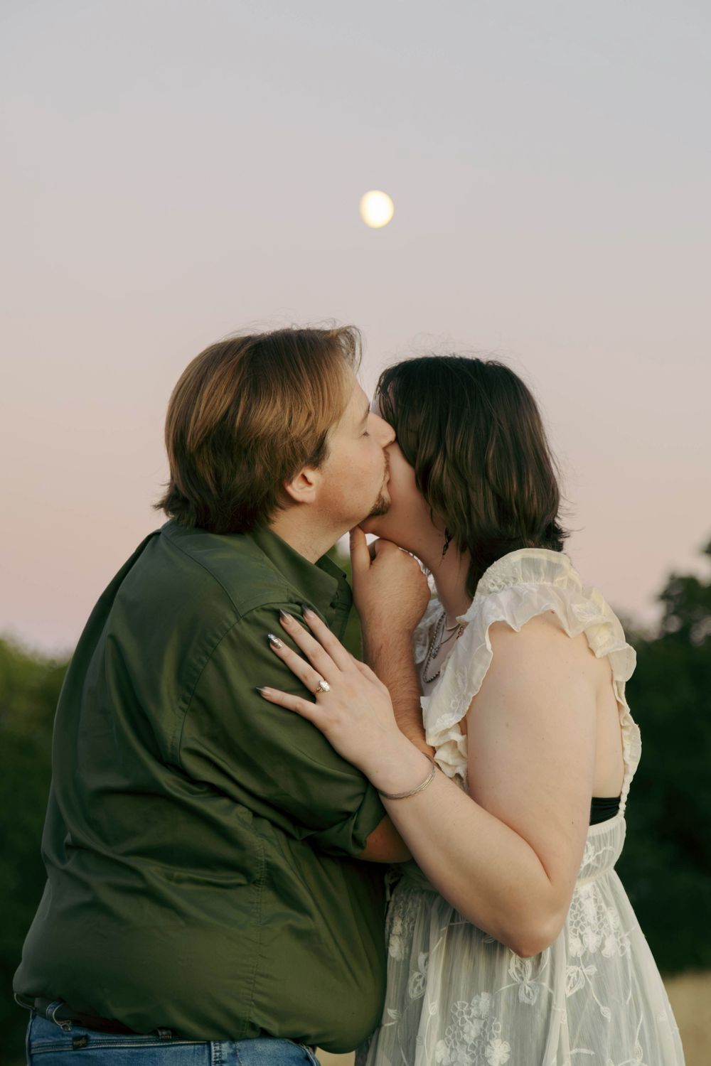 Couple sharing a romantic kiss outdoors at dusk with a full moon glowing in the pastel sky behind them.