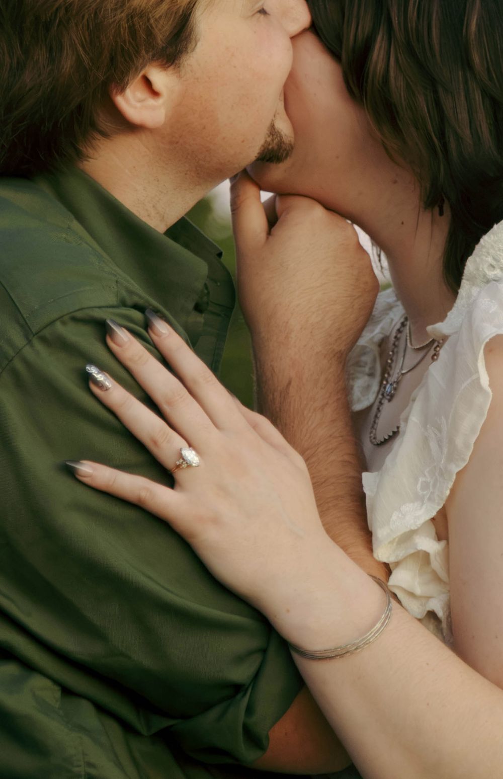 Couple sharing a romantic kiss, woman wearing engagement ring and bracelet, man in green shirt.
