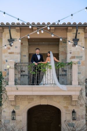 A couple stands on separate balconies of a stone building with string lights and lanterns decorating the exterior.
