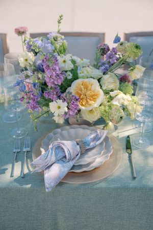 Elegant table setting with pastel floral centerpieces featuring lilac, peach roses and greenery on a sage green tablecloth.