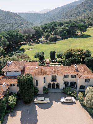 Aerial panoramic view of Mediterranean style homes with terracotta roofs nestled in green hills with mountains in background.