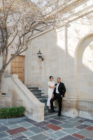A couple poses playfully on stone steps against a white stone building with arched architecture.