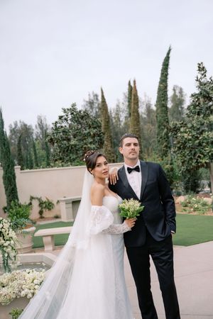 Bride in white lace gown with veil and groom in black tuxedo pose together in elegant garden wedding venue.