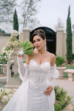 Bride in white lace off-shoulder gown holds green and white bouquet outdoors near elegant stone entrance.