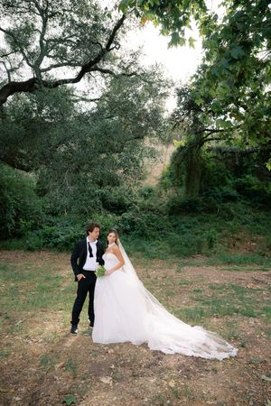 Bride in flowing white gown and groom in black tuxedo pose together in a lush green outdoor garden setting.