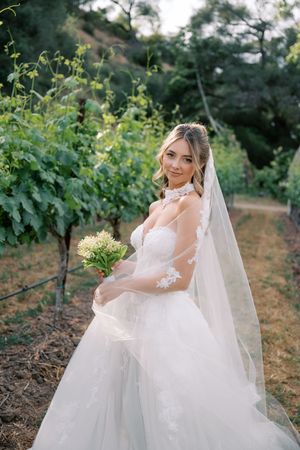 Bride in lace wedding gown and veil holds bouquet among lush vineyard rows on her wedding day.