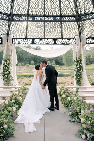 Bride and groom share first kiss under ornate iron gazebo adorned with white florals and greenery.