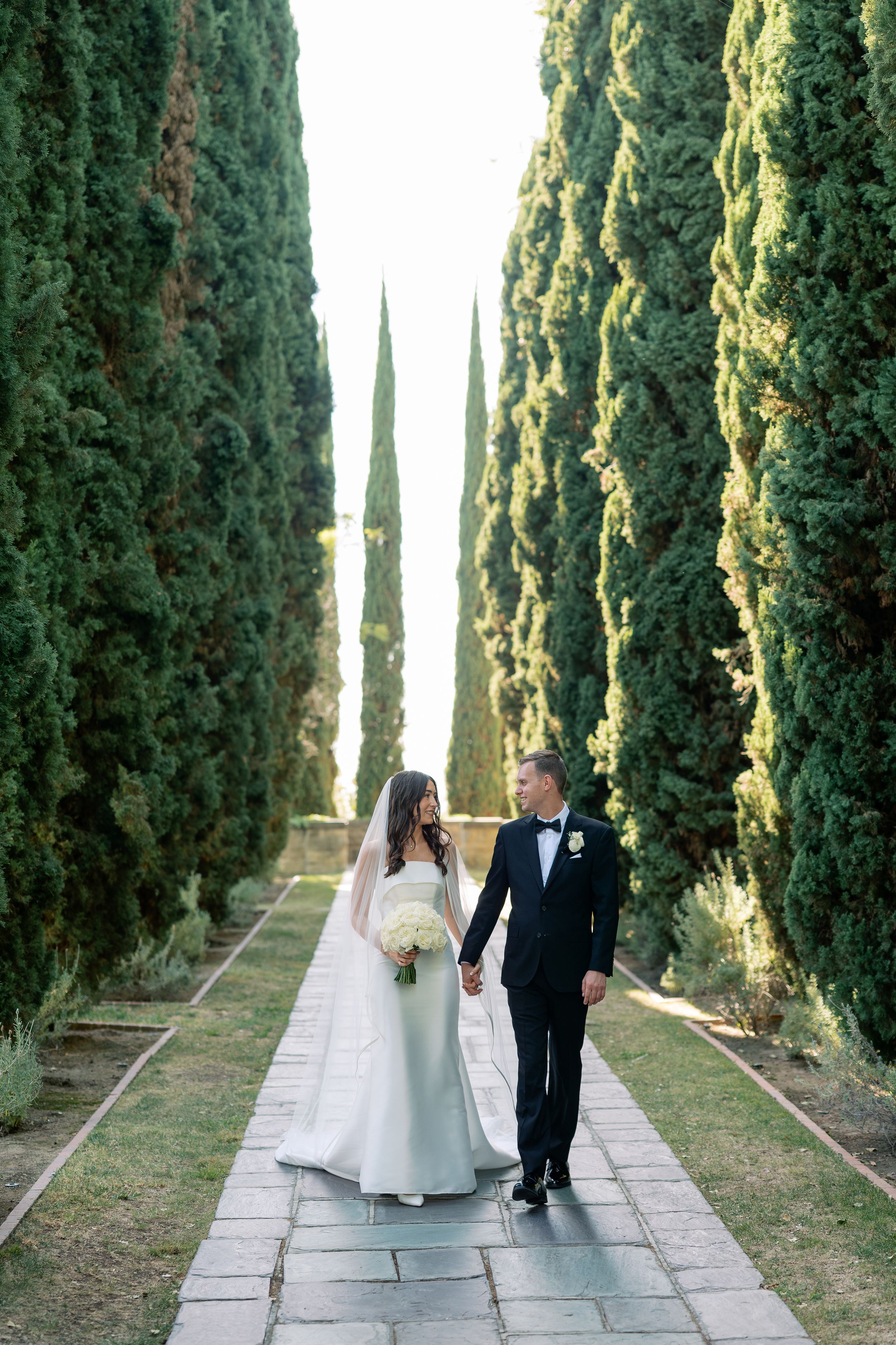 Bride in a white gown holding a bouquet walks hand in hand with groom in a dark suit along a stone path lined with tall cypress trees.