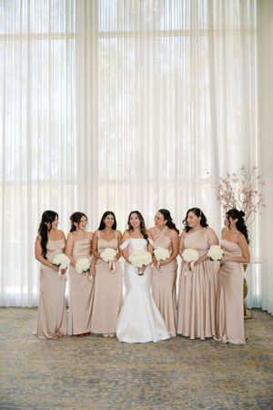 Bride in white gown stands with six bridesmaids in champagne dresses holding white bouquets before sheer curtains.