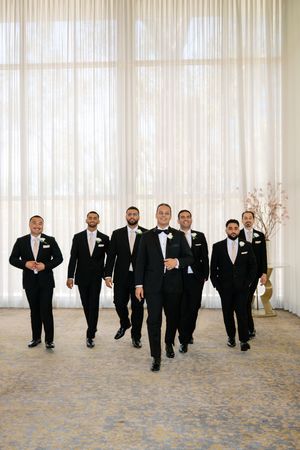 Groom leads groomsmen in black tuxedos walking confidently in elegant venue with sheer curtains.
