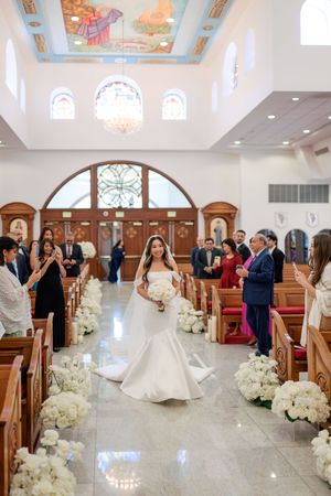 Bride walks down the aisle of a decorated church, holding white bouquet, with guests standing in wooden pews.