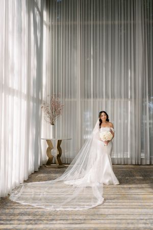 Bride in fitted white gown with long cathedral veil holds bouquet in sunlit ballroom.