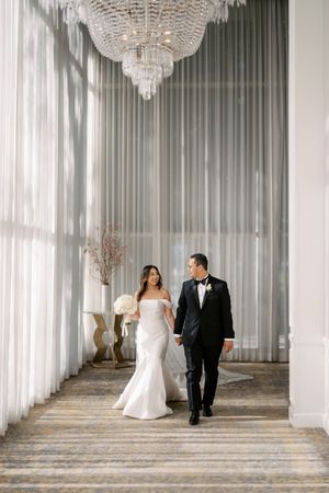 Elegant bride and groom hold hands walking under crystal chandelier in luxury hotel corridor.