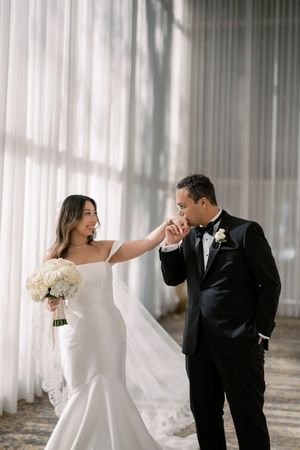 Groom kisses bride's hand as she smiles holding white roses bouquet in elegant venue.