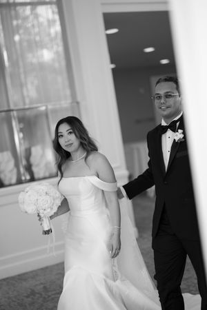 Black and white photo of bride in off-shoulder gown holding bouquet with groom in tuxedo at wedding venue.