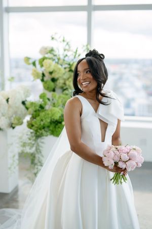 Bride in a stunning white ball gown holds pink roses while posing near floral arrangements with city views.