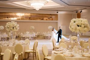 Bride and groom share first dance in elegant white ballroom with tall floral centerpieces and chandelier lighting.