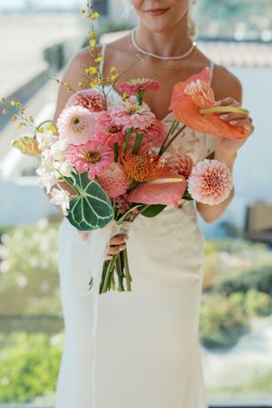 Elegant coral and pink dahlia wedding bouquet held against a white dress in natural lighting.