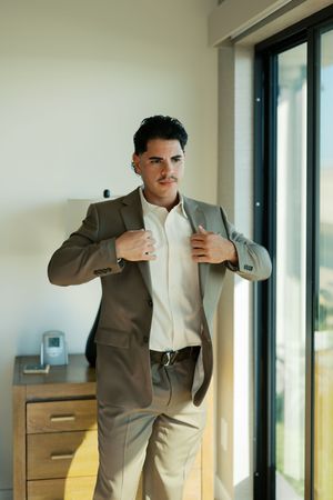 A person in a light gray suit adjusts their jacket while standing near large windows in a bright room.