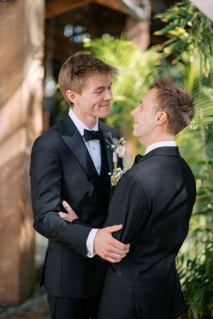 Two grooms in black tuxedos share a tender moment, smiling at each other outdoors amid lush tropical greenery.