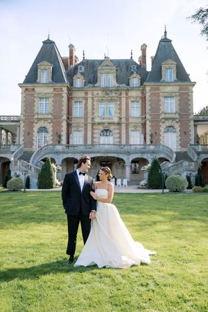 Bride and groom pose on lush lawn before elegant French château wedding venue on sunny day.