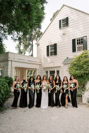 Bride in white lace gown poses with eight bridesmaids in black dresses holding bouquets outside elegant white estate.