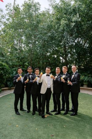 Groom in white tuxedo jacket raises champagne glass with six groomsmen in black suits outdoors.
