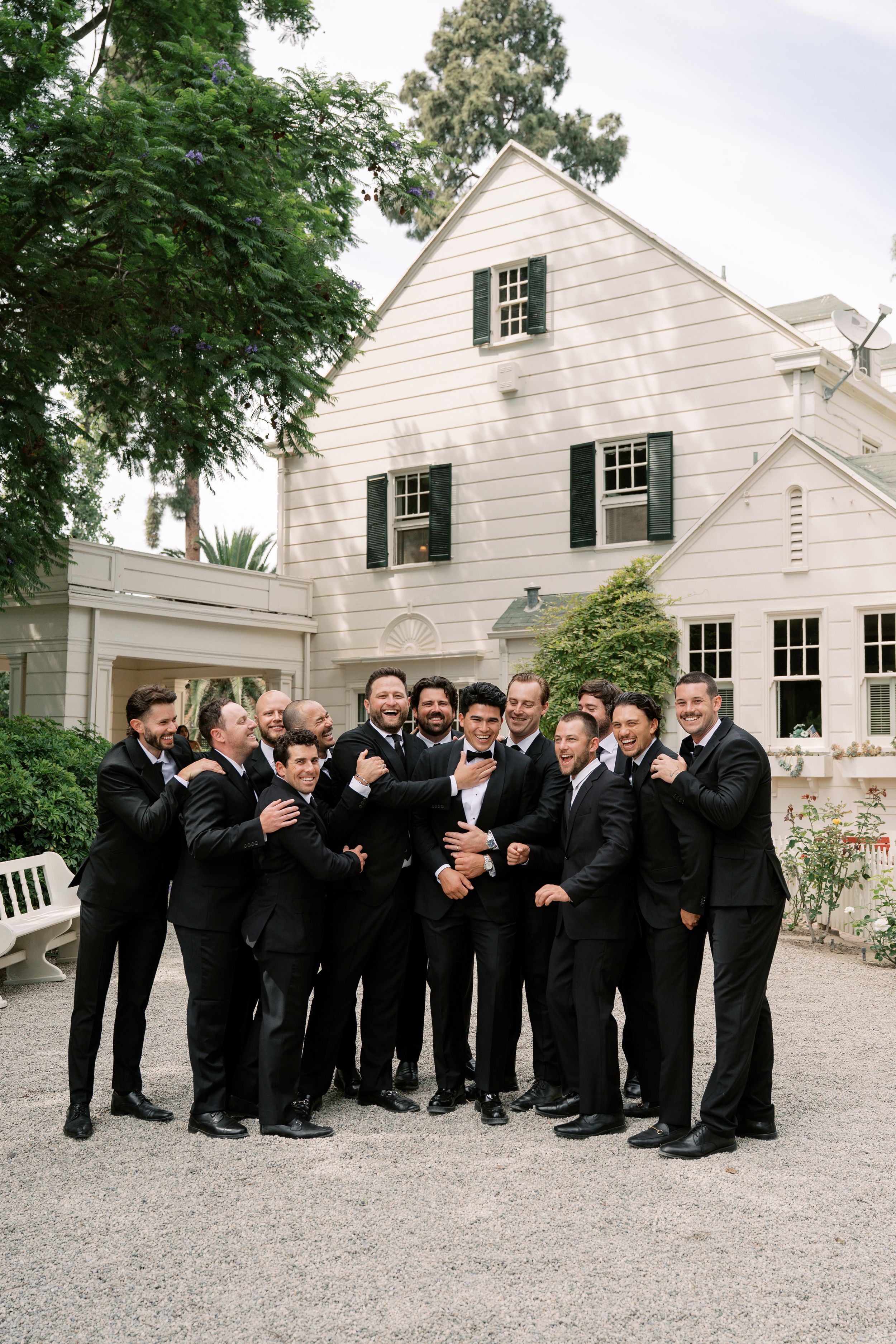Groom and groomsmen in black tuxedos laughing together outside a white colonial-style wedding venue.