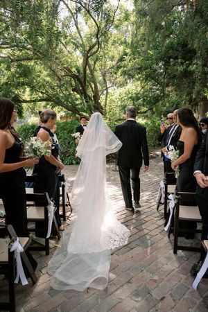 Bride walking down outdoor aisle with father, long white veil trailing, guests in black attire seated among lush green trees.