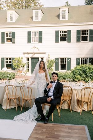 Bride in lace gown and groom in tuxedo pose at outdoor wedding reception beside a white colonial estate.