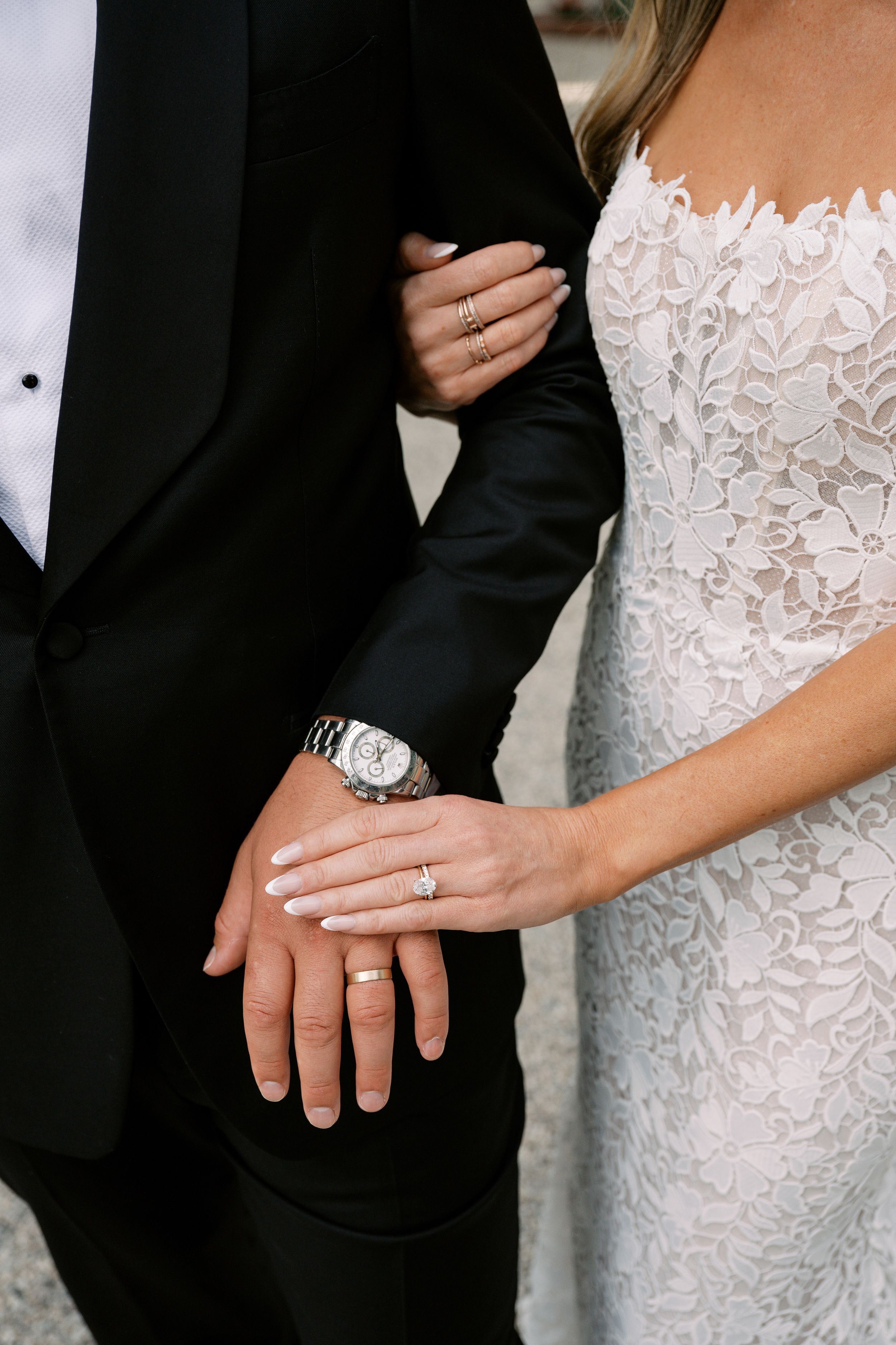 Bride and groom displaying wedding rings, bride in lace gown, groom in black tuxedo, close-up of hands.